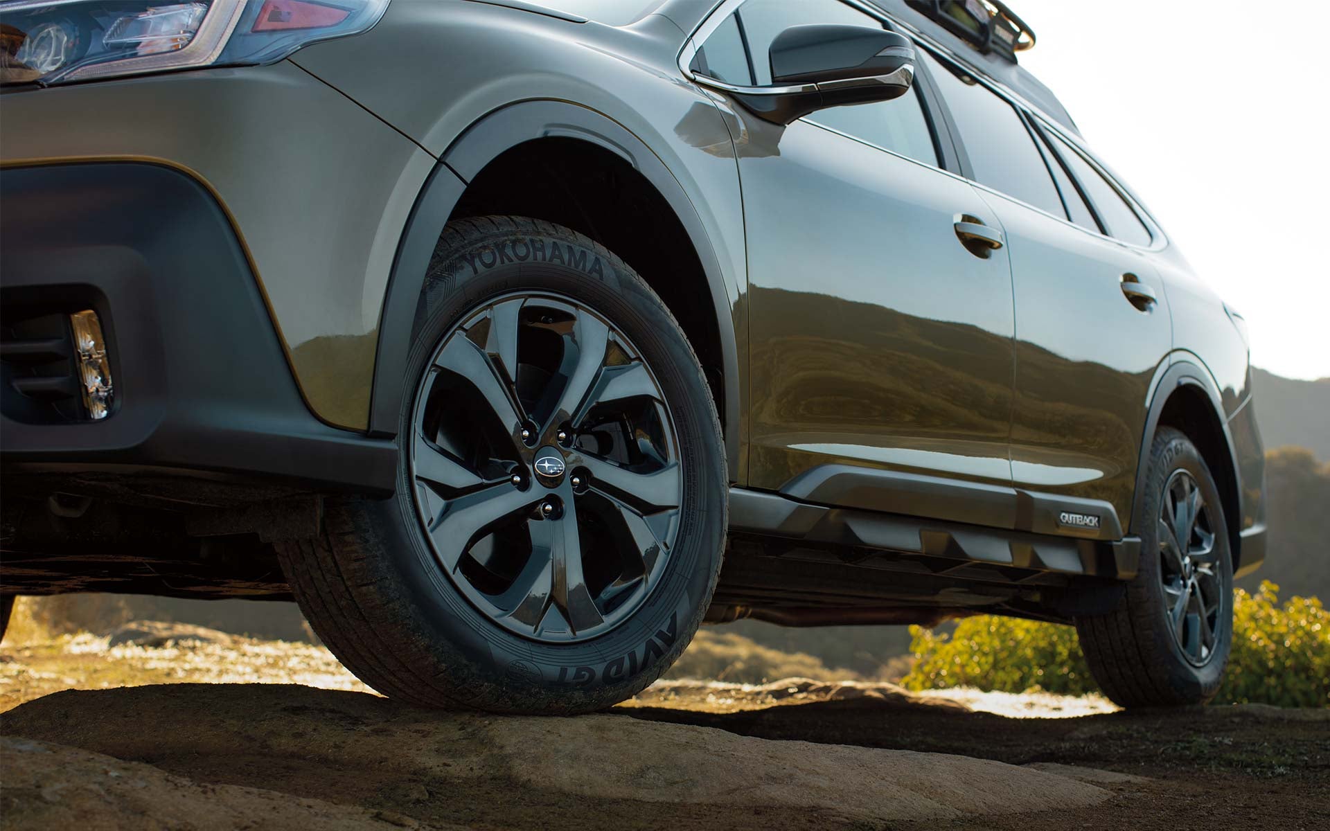 Close-up view of a Subaru Outback's front wheel, tire, and lower body panels positioned on rugged rocky terrain